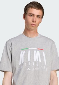 Grå bomulds t-shirt med korte ærmer, der har et iøjnefaldende hvidt print "KIMI" og grønne og røde accenter. Inkluderer Adidas og AMG Petronas logoer.