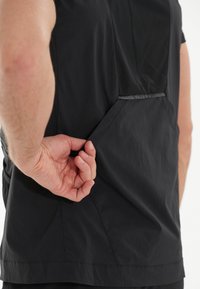 Svart idrottströja med en nätpanel på ryggen, mjuk yta och en diskret sidficka. Starkt, lättviktigt tyg.