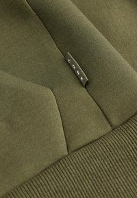 Felpa verde oliva con texture morbida, caratterizzata da polsini e orlo a coste, e un piccolo etichetta "CORE" cucita di lato.