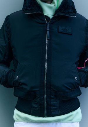 Personne portant une veste bomber noire zippée avec un patch sur la poitrine et les mains dans les poches, par-dessus un sweat à capuche vert clair.