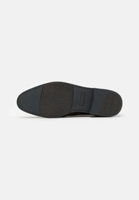 Semelle de chaussure noire en caoutchouc texturé, avec une zone de nez lisse et une section de talon offrant du soutien. Logo embossé sur le talon.