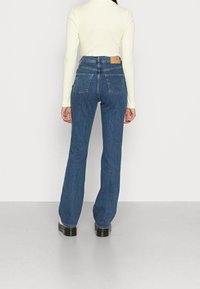 Jeans de denim azul de cintura alta y acampanados con un ligero estiramiento, que cuentan con dos bolsillos traseros y una etiqueta de marca de cuero marrón en la cintura.