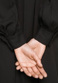 Vêtement noir à manches bouffantes, tissu texturé, et boutons décoratifs aux poignets ; les mains sont jointes derrière le dos.