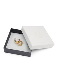 Guldtonad smycken visade i en grå sammetspäckad låda med ett vitt strukturerat lock som har präglad logotyp i guld.