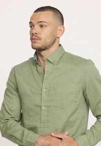 Chemise verte à boutons avec une poche poitrine, une texture subtile et un col doublé contrastant. Coutures élégantes et boutons ronds.