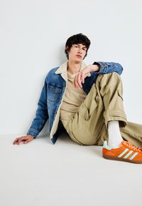 Jeansjacke mit Fellkragen, beiges T-Shirt, Cargo-Hosen und orangefarbene Sneakers mit tealfarbenen Akzenten. Modell sitzt entspannt.