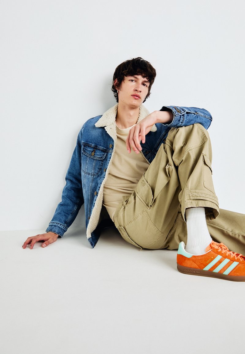 Chaqueta de mezclilla con cuello de piel, camiseta beige, pantalones cargo y zapatillas naranjas con acentos en azul verdoso. Modelo sentado con postura relajada.