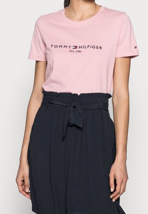 Nainen pukeutunut vaaleanpunaiseen Tommy Hilfiger -T-paitaan, jossa logo, ja mustaan korkeavyötäröiseen hameeseen, joka on solmittu vyötärölle.