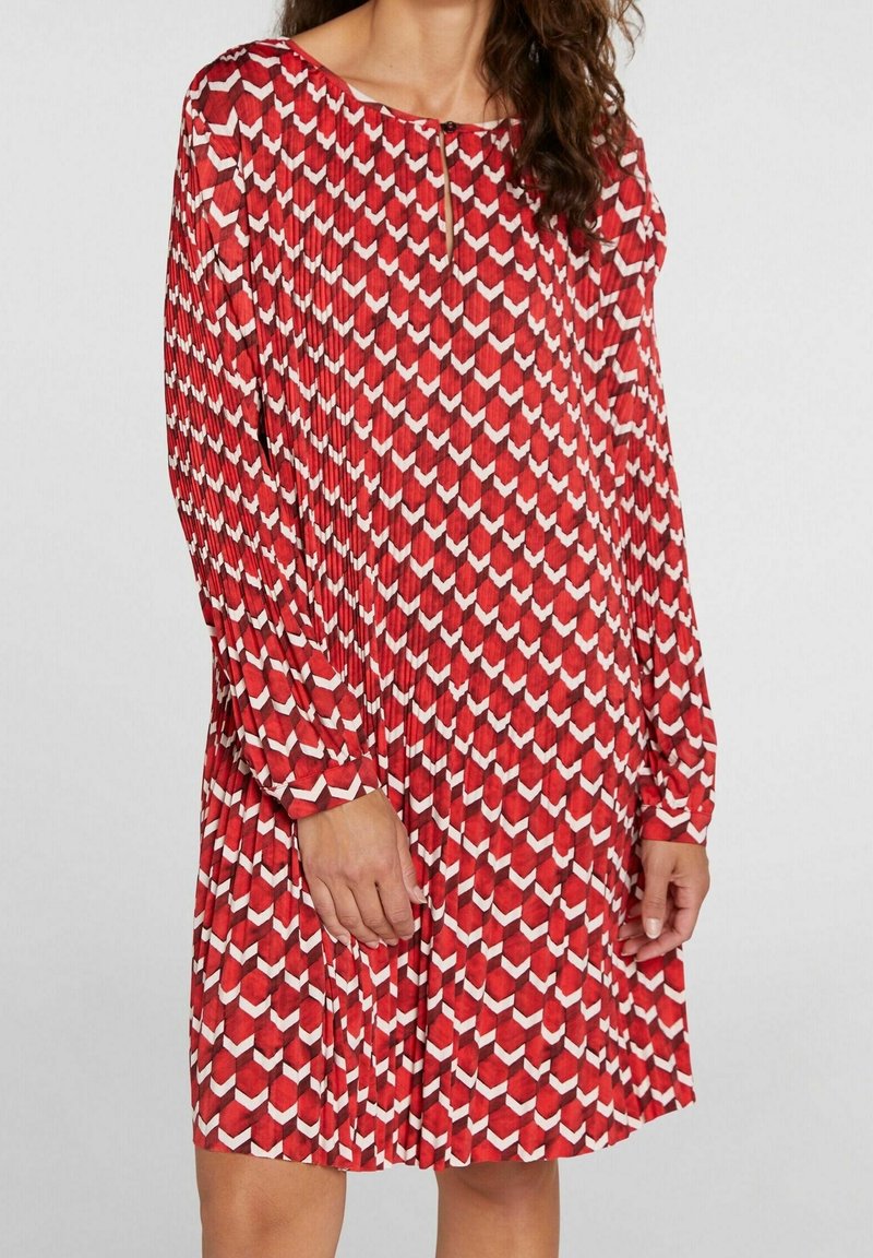 Robe plissée rouge avec un motif géométrique blanc, manches longues, encolure ronde et un détail en trou de serrure à l'avant.