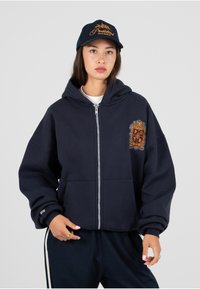 Marineblå hettegenser med glidelås, frontlomme og stort brodert logo i gull og rødt. Har en romslig hette og ribbestrikkede mansjetter.