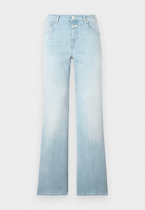 Femme aux longs cheveux foncés portant un T-shirt bleu clair, un jean clair avec une ceinture noire et des sandales noires à talons, debout, les mains dans le dos.