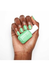 Vernis à ongles vert dans un flacon carré étiqueté "essie", tenu dans une main avec des ongles soigneusement manucurés peints de la même teinte de vert.