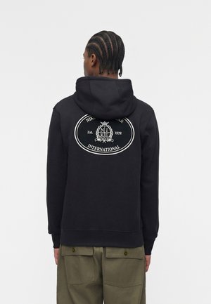 Homme aux cheveux tressés portant un sweat à capuche Nike noir avec un logo circulaire blanc et un texte dans le dos, associé à un pantalon vert olive, de dos.