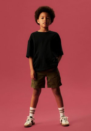 Bērns ar cirtainiem matiem stāv priekšā rozā fonam, valkājot melnu T-kreklu, olīvu krāsas kargo šortus, svītrainas zeķes un baltas sporta kurpes.