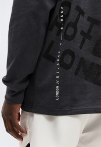 Κοντινό πλάνο ενός ατόμου που φοράει μαύρο μακρυμάνικο πουκάμισο με το κείμενο «LONDON //51.6042 N 0.0662» και εκρού παντελόνι.