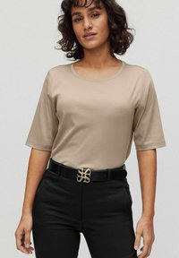 Beige t-shirt med kort ärm och rund hals, i kombination med svarta byxor och en dekorativ guldspänne med ett stiliserat mönster.