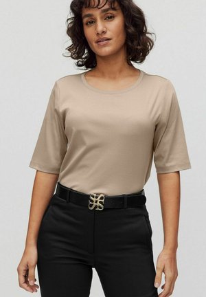 Beige short-sleeve t-shirt with a round neck, paired with black pants and a decorative gold belt buckle showing a stylized design.