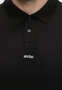 Prim-plan al gulerului unui tricou polo negru cu doi nasturi și logo-ul alb „HUGO” centrat sub fenta nasturilor.