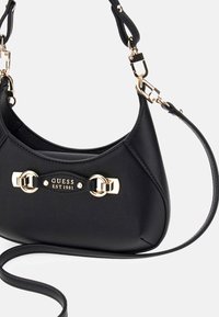 Bolso de mano negro con acabado texturizado, acentos en hardware dorado y una placa con el logo. Presenta una sola correa para el hombro y un diseño curvado.