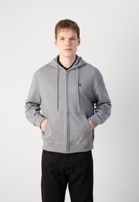 Grijze zip-up hoodie van zacht materiaal, met een voorvak, een capuchon met trekkoord en een geborduurd logo op de borst. Zwarte broek eronder.