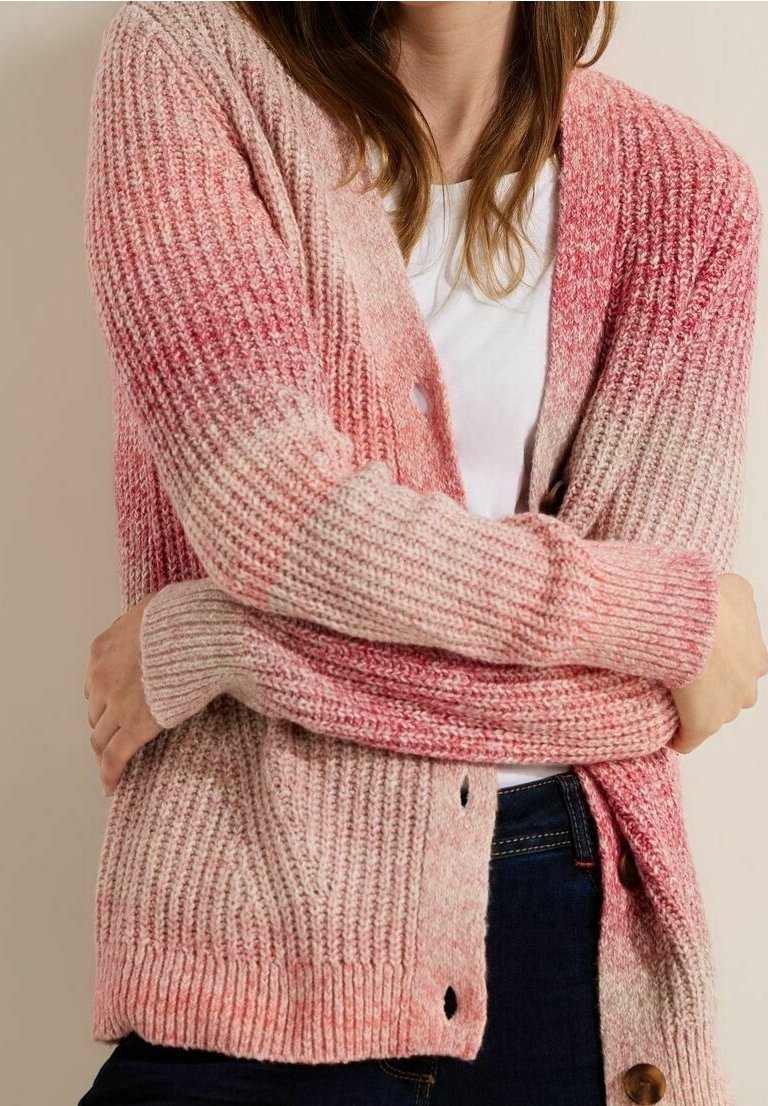 Femme portant un cardigan en maille côtelée rose et beige sur une chemise blanche, les bras croisés, jean foncé visible.