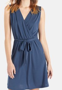 Femme portant une robe portefeuille bleu foncé sans manches avec ceinture nouée à la taille sur fond blanc.