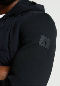 Crni atletski prsluk s gornjim dijelom od najlona i rukavima od mješavine pamuka. Značajni detalji uključuju gumenu oznaku na rukavu i podešavanja kapuljače.