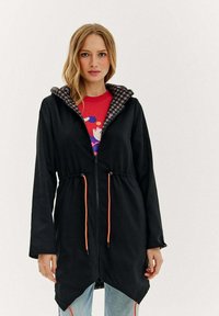 Sieviete ar melnu jaku ar rāvējslēdzēju, kurai ir pārbaudīta kapucnes odere, oranžām auklām, sarkanu rakstainu kreklu un gaiši ziliem džinsiem.