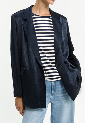 Person trägt ein glänzendes dunkelblaues Sakko über einem weiß-navy gestreiften Hemd mit hellblauen Jeans, Hände in den Taschen.