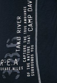 Camiseta azul marino con texto y gráficos en blanco. Presenta cuello redondo, mangas cortas y costuras decorativas a lo largo del costado.