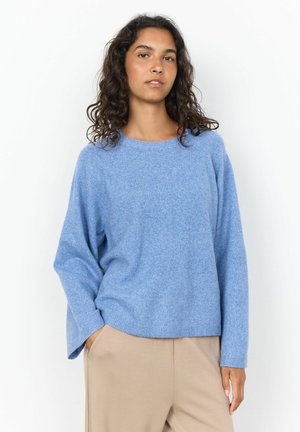 Femme aux cheveux bouclés portant un pull ample bleu clair et un pantalon beige, debout avec une main dans la poche sur un fond blanc.
