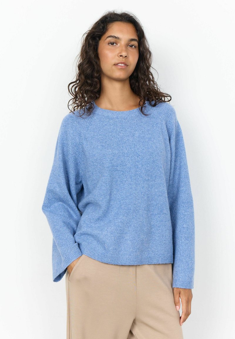 Femme aux cheveux bouclés portant un pull ample bleu clair et un pantalon beige, debout avec une main dans la poche sur un fond blanc.