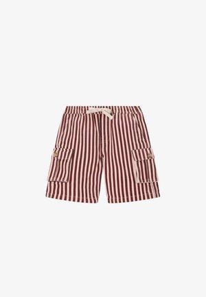 Garçon aux cheveux bouclés portant un t-shirt jaune, un short rayé rouge et blanc, des baskets, et portant un sac à dos vert à motifs sur une épaule.