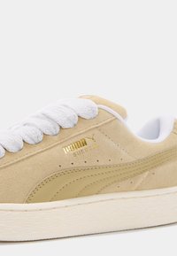 La sneaker Puma en suède beige présente une tige texturée, des lacets blancs, un logo doré et une semelle en caoutchouc avec un motif ondulé.