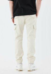 Beige cargobukser med sidelommer, elastiske manchetter og en sort detalje, parret med hvide sneakers. Enkel design og blødt materiale.