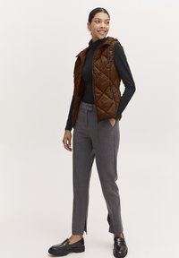 Gilet matelassé marron avec capuche, comprenant des poches avant et une fermeture éclair, associé à un pantalon gris ajusté et des mocassins noirs.