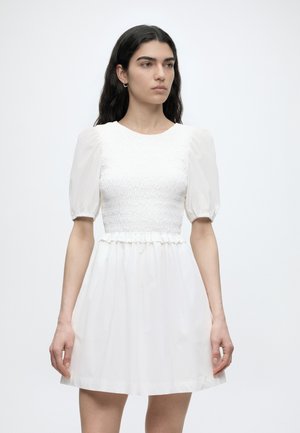 Vrouw met lang donker haar, die een witte korte mouw jurk met opgeblazen mouwen en een geplooide taille draagt, staand tegen een effen achtergrond.