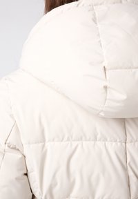 Lichtbeige pufferjack met een capuchon, voorzien van een rechthoekig gequilt patroon en een gladde, glanzende textuur. Gestikte naden zijn zichtbaar op de schouder.