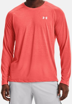 Mężczyzna w koralowej, długorękawowej sportowej koszulce z białym logo Under Armour i jasnoszarych spodenkach, stojący na jednolitym tle.