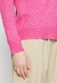 Rosa stickad tröja med ribbad muddar och en något fransig fåll, parat med ljusbeiga byxor. Mjuk textur och avslappnad passform.