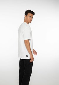 Witte t-shirt met korte mouwen en een ontspannen pasvorm, gemaakt van katoen, met een klein zwart labelaccent aan de onderkant van de zoom. G gedragen met zwarte broek.