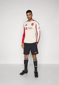 Rotes und cremefarbenes Fußballtrikot mit langen Ärmeln und Rundhalsausschnitt, mit Logo und roten Streifen, kombiniert mit schwarzen Shorts und Socken.