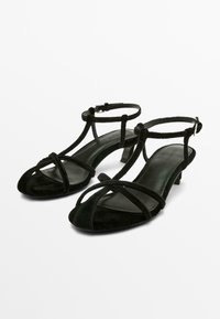 Sandales noires à talon bas avec des fines brides en velours qui se croisent sur les orteils et se ferment autour de la cheville avec de petites boucles.