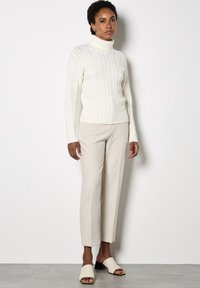 Maglione a collo alto in maglia a trecce di colore crema, pantaloni beigi a gamba affusolata e mules chiare, che mettono in risalto dettagli texturizzati e una silhouette snella.