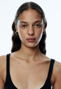 Zwarte geribbelde tanktop met brede banden, met een textuurpatroon. Huidkleur is licht, met natuurlijke make-up en kleine ringoorbellen.