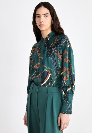 Grün gemusterte Bluse mit Kettenmotiven in Braun- und Goldtönen, mit knopflosem Frontverschluss und langen Ärmeln. Kombiniert mit grünen maßgeschneiderten Hosen.
