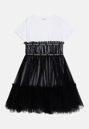 Robe blanche à manches courtes avec jupe en tulle noir superposée et deux bandes noires avec texte blanc "GUESS" sur la poitrine et la taille.