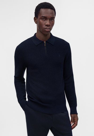 Jeune homme portant un pull bleu marine texturé à manches longues avec un col zippé, debout, une main dans la poche.