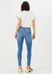 Vaqueros ajustados de color azul con una cintura de altura media, que cuentan con bolsillos traseros y una etiqueta de la marca. Se llevan con una camiseta blanca de manga corta y sandalias de plataforma marrones.