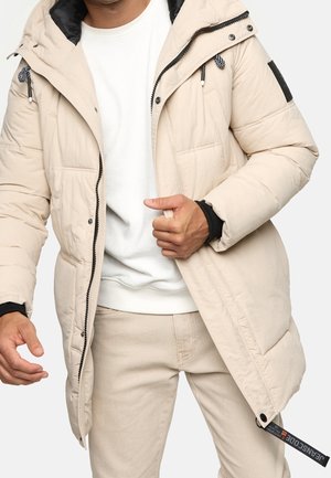 Mann trägt beige gesteppte Jacke mit schwarzen Reißverschluss-Riegeln, weißem Sweatshirt und passenden beigen Hosen, richtet die Jackenvorderseite aus.